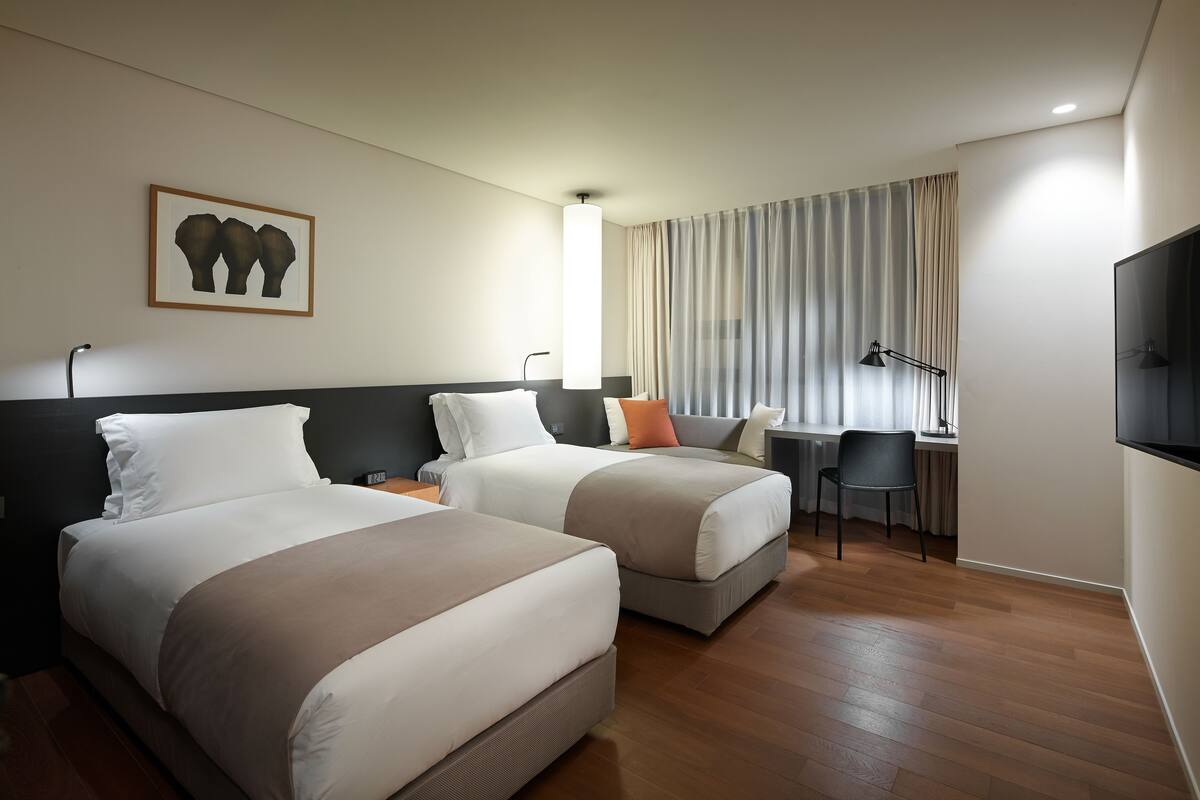 A hotel room with two beds, a desk, a chair, a TV, and a window with curtains.