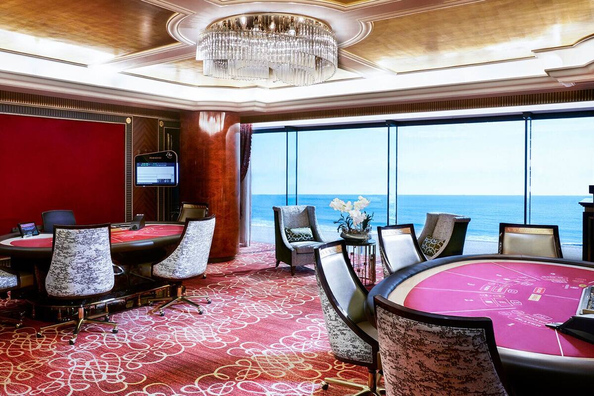 A casino room with a large roulette table, multiple gaming tables, and a view of the ocean.