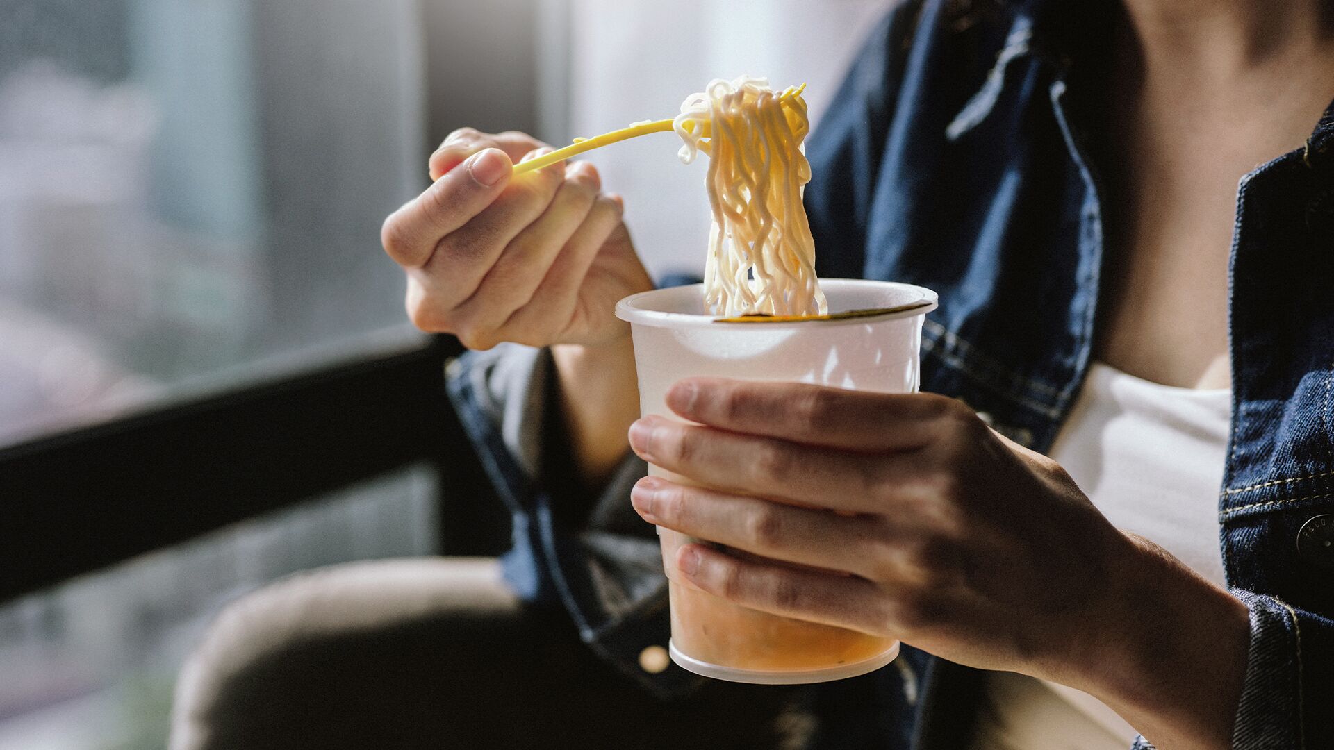 A person holding a cup with noodles and a yellow spoon.
