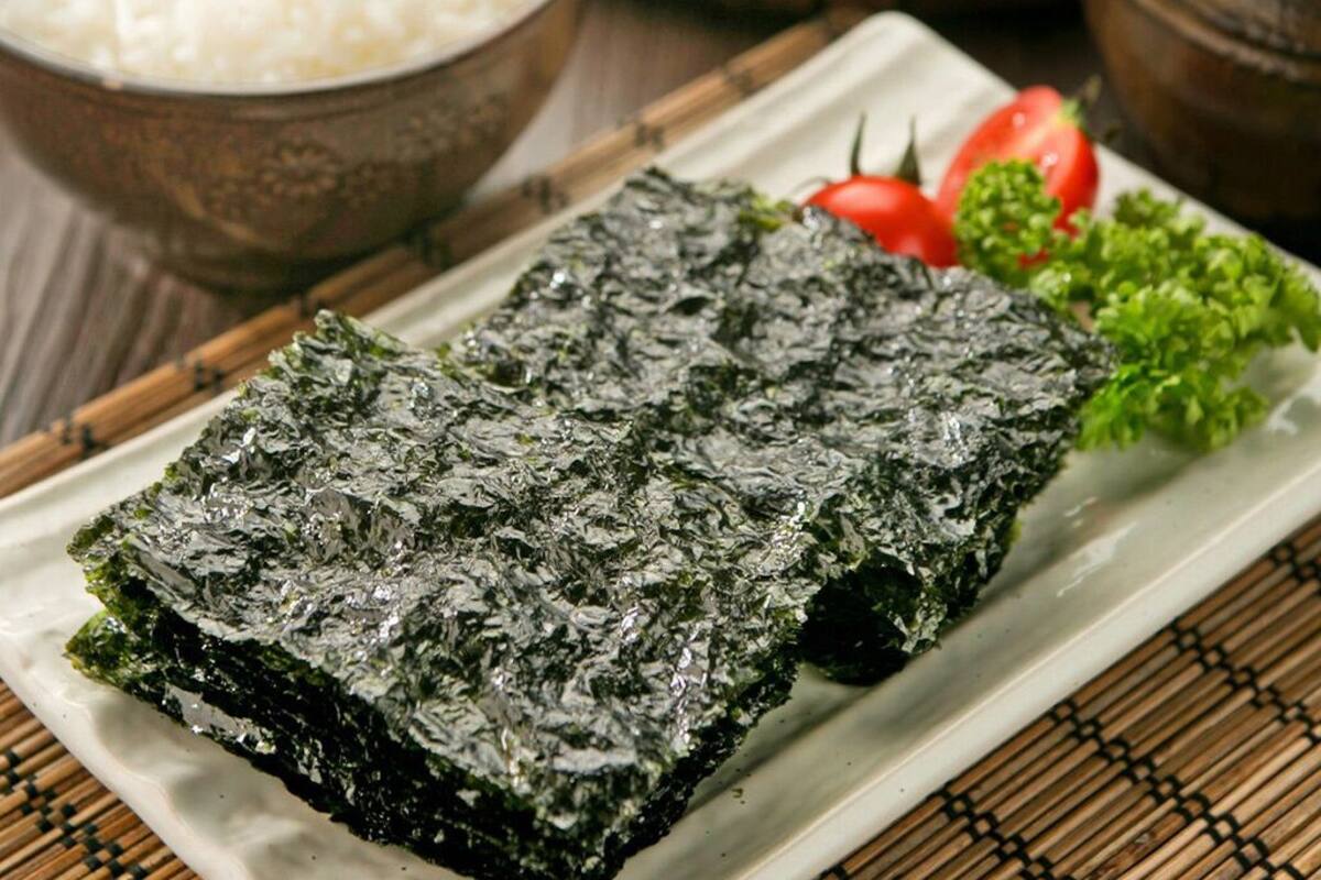 A rectangular plate with a dark green seaweed dish, a small bowl of white rice, a tomato, and some green lettuce on a bamboo mat.