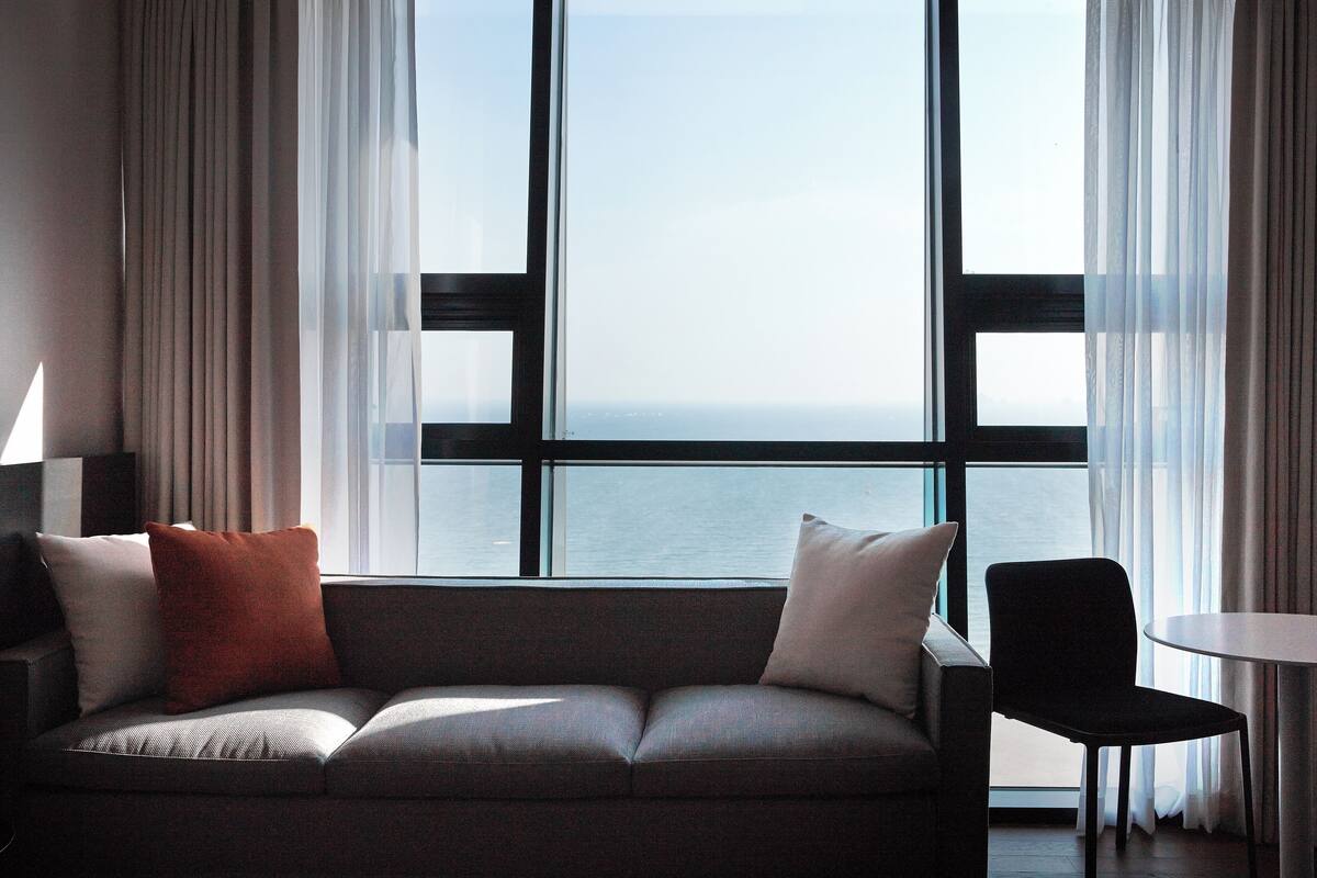 A modern living room with a sofa, a chair, and a small table. A large window offers a view of the sea.