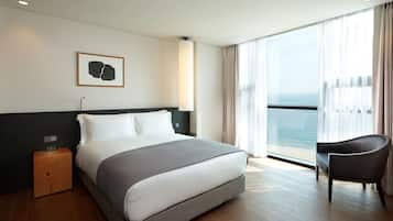 Una moderna camera d'albergo con un letto grande, un tavolino, una poltrona e vista mare attraverso ampie finestre.