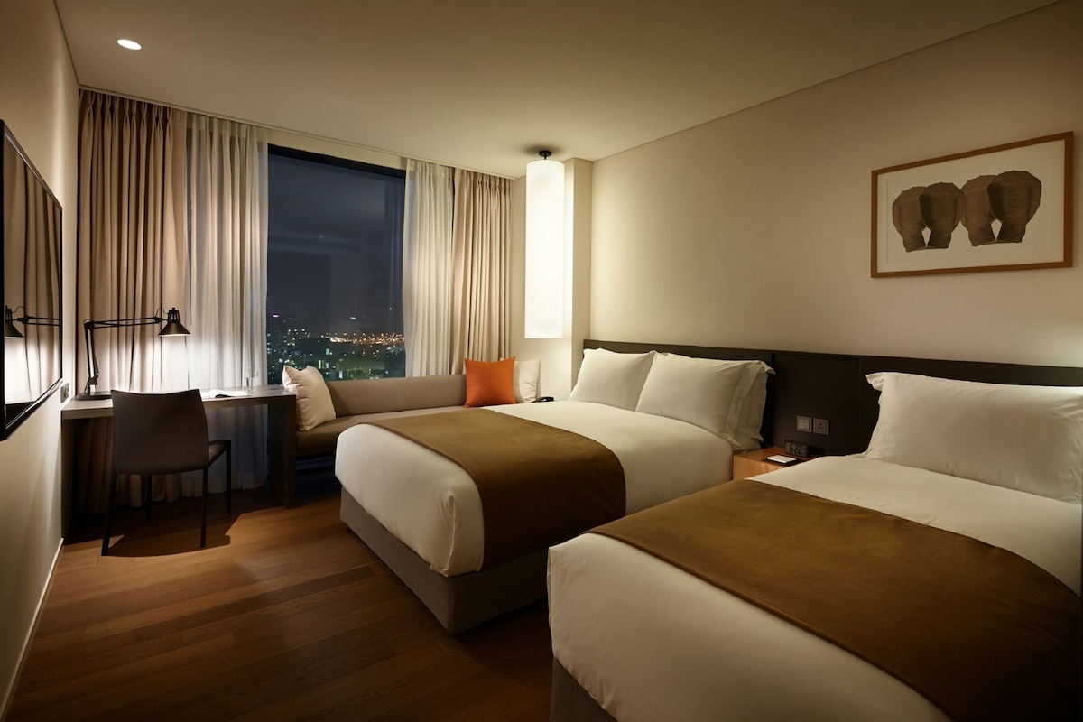 Habitación de hotel con dos camas, un escritorio, una silla y un ventanal con vistas a la ciudad.
