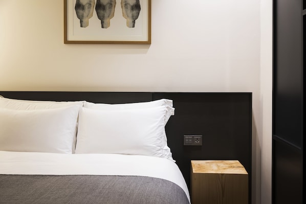 A neatly made bed with white linens, a dark headboard, and a small wooden nightstand beside it.