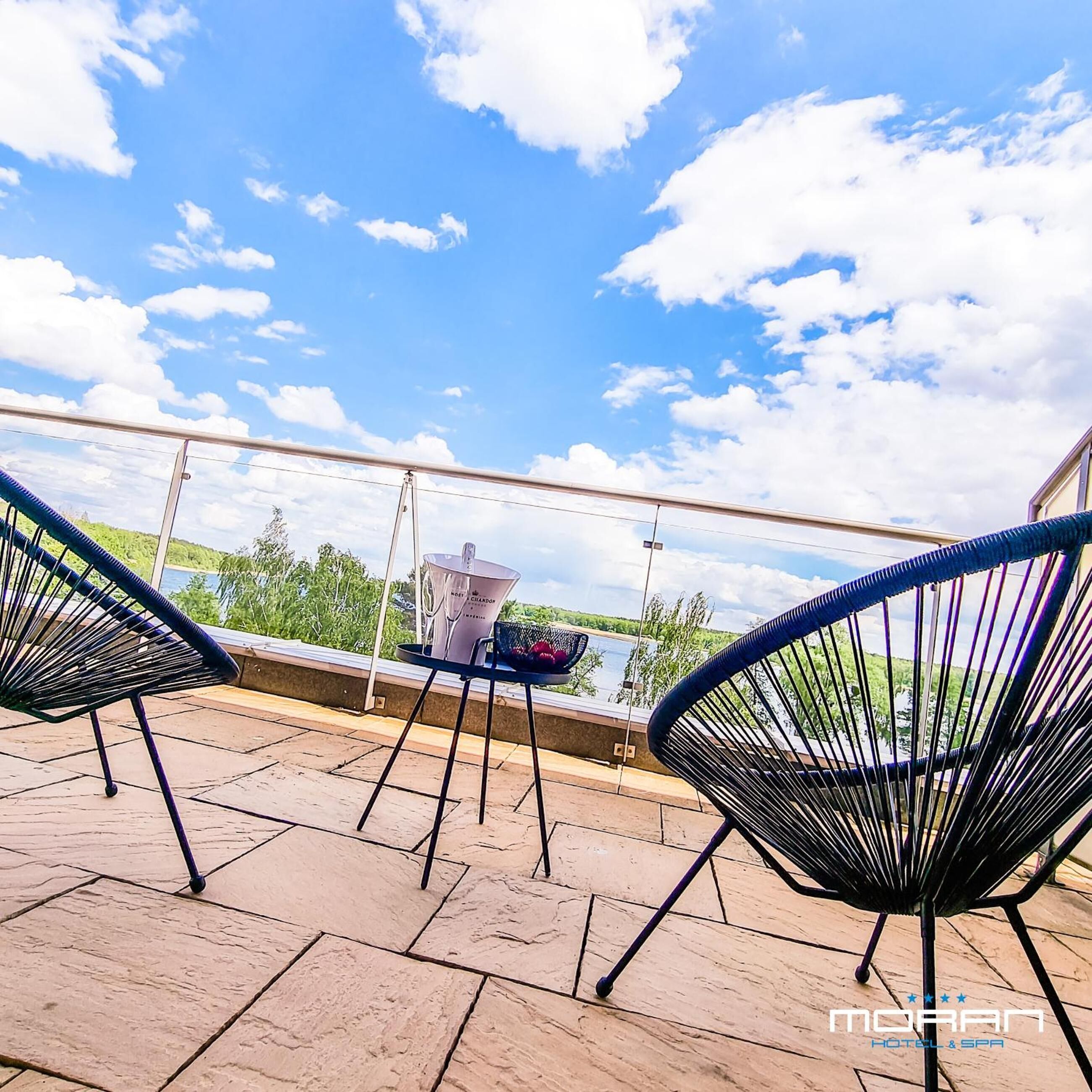 Une terrasse avec deux chaises à l’armature en fil de fer noir, une table sur laquelle se trouve un pichet blanc, et une vue sur un lac et la végétation, sous un ciel partiellement nuageux.