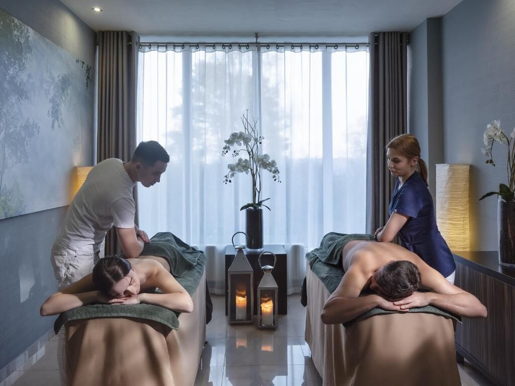 Un cuarto de masajes con dos clientes recibiendo tratamiento, un terapeuta atendiendo a un cliente y un jarrón con flores sobre una mesa.