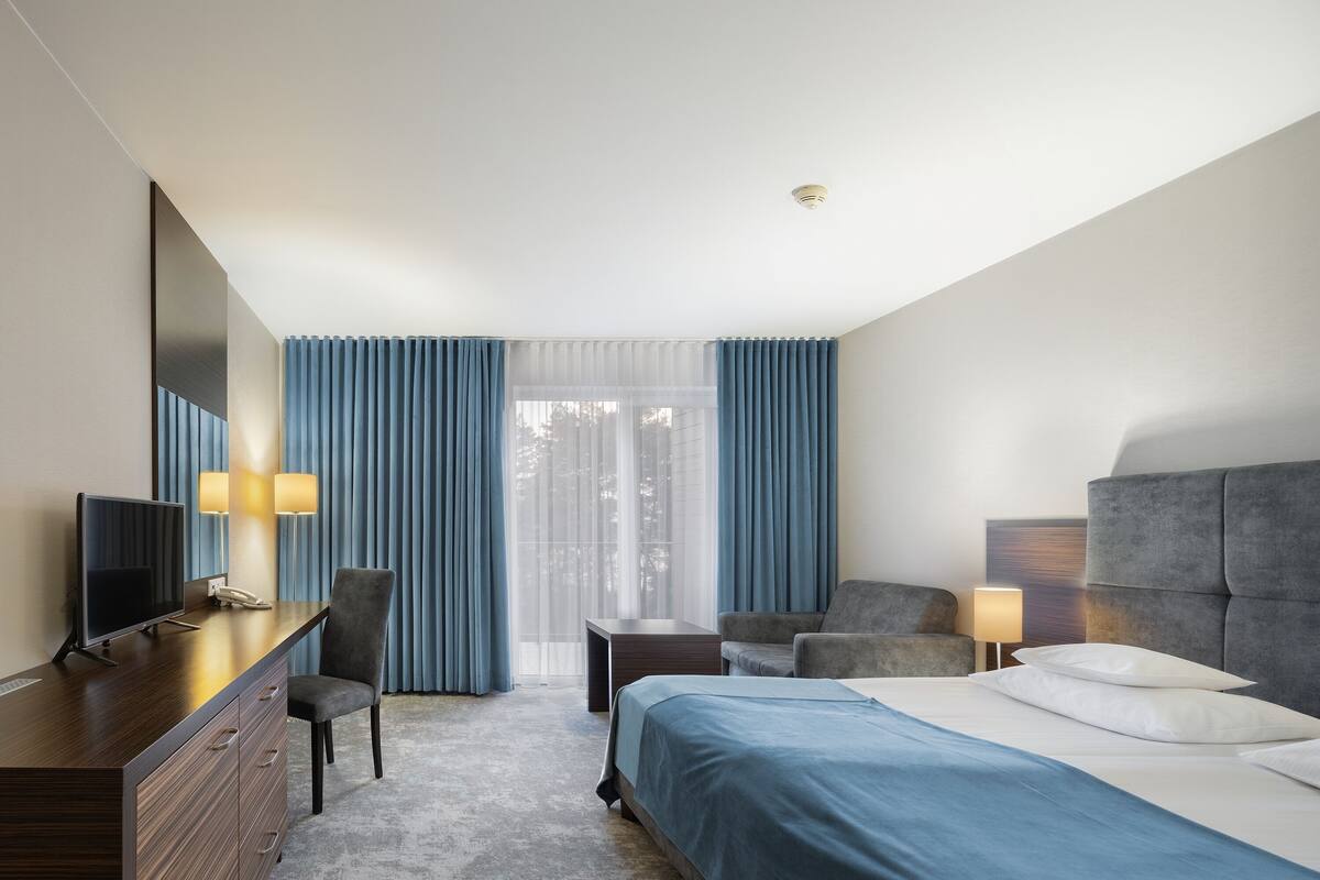 A hotel room with a bed, a desk with a computer, a chair, a small table, and a television.