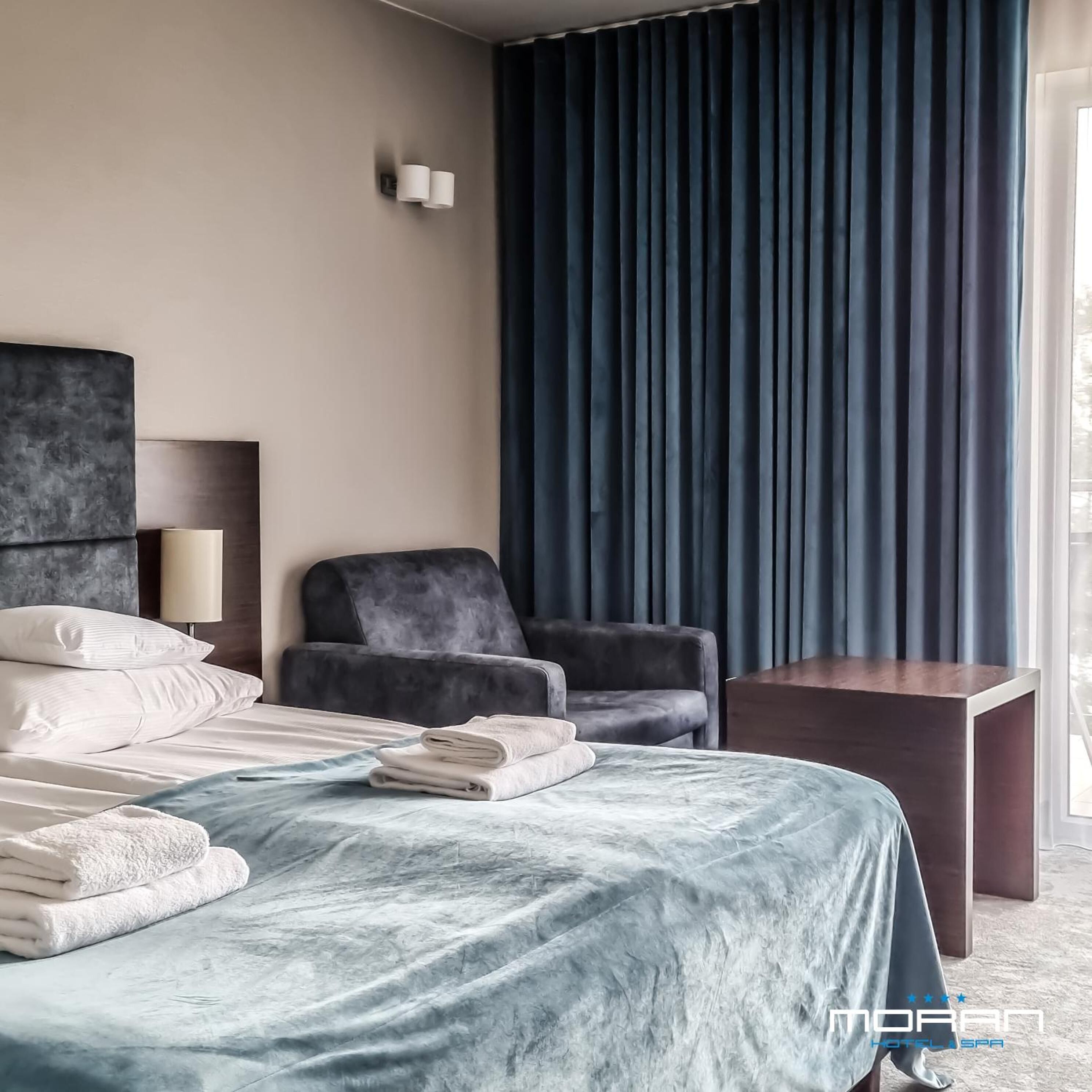 Habitación de hotel moderna con cama, una silla, una mesita de noche y un ventanal con cortinas oscuras.