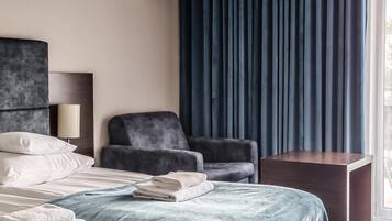 Habitación de hotel moderna con cama, una silla, una mesita de noche y un ventanal con cortinas oscuras.
