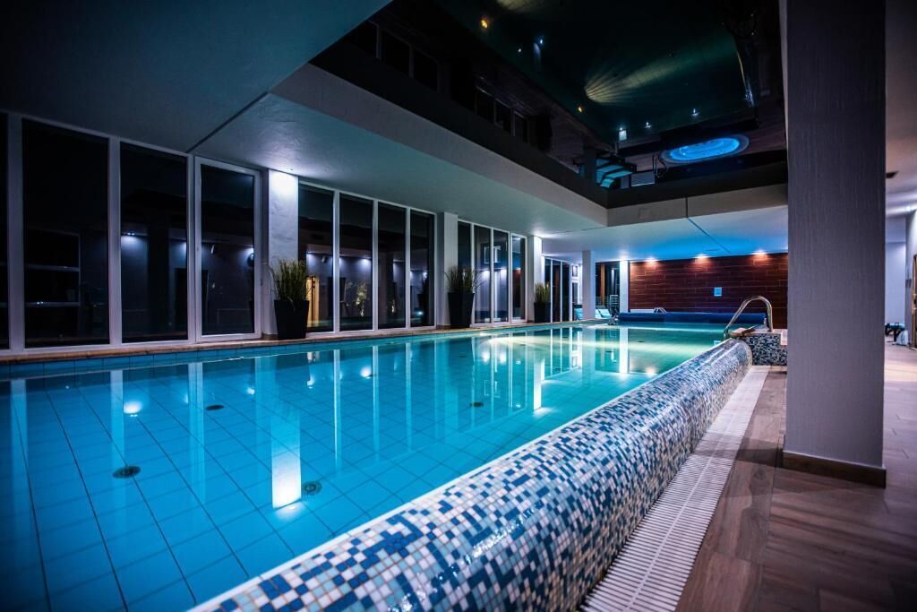 An indoor swimming pool with clear water, surrounded by glass walls and wooden flooring.