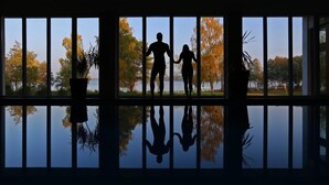 Silueta de una pareja de pie junto a una ventana con vista a árboles y una masa de agua.