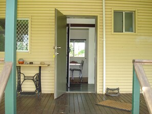 A wooden porch with a metal railing, a small table with a sewing machine, and a door leading to an interior space with a chair and a table.