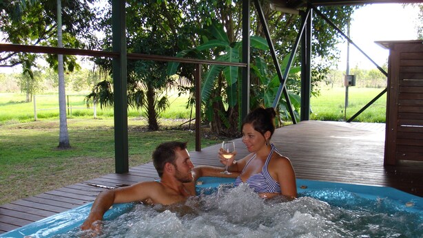 Un hombre y una mujer en un jacuzzi con una terraza de madera y follaje en el fondo.
