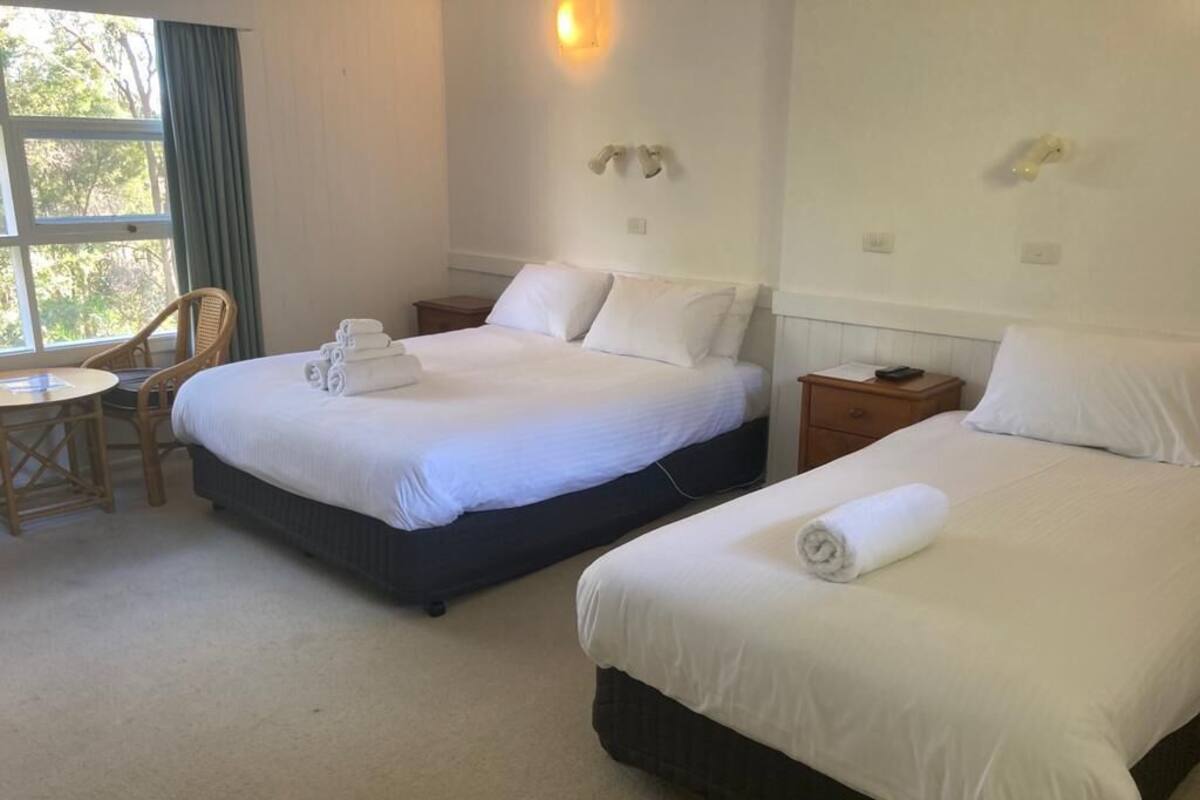 Two beds with white linens, a small round table, and a chair in a room with a window and wall-mounted light fixtures.