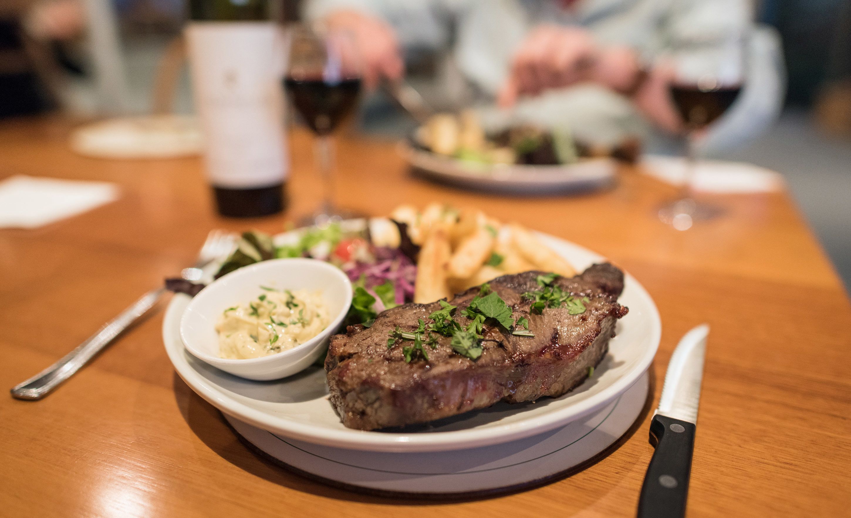 A plate of steak with a side of fries and a small bowl of sauce on a wooden table.