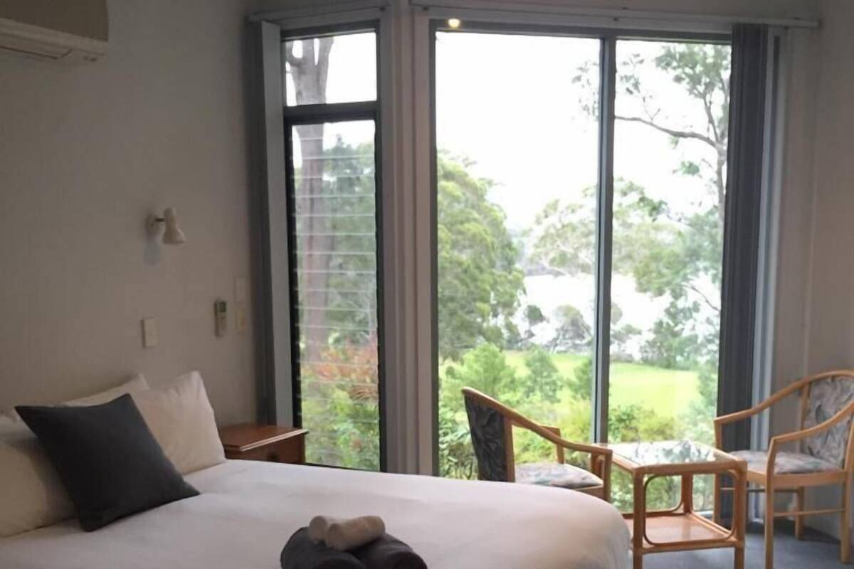 A bedroom with a large bed, a ceiling fan, and a view of the outdoors through large windows.