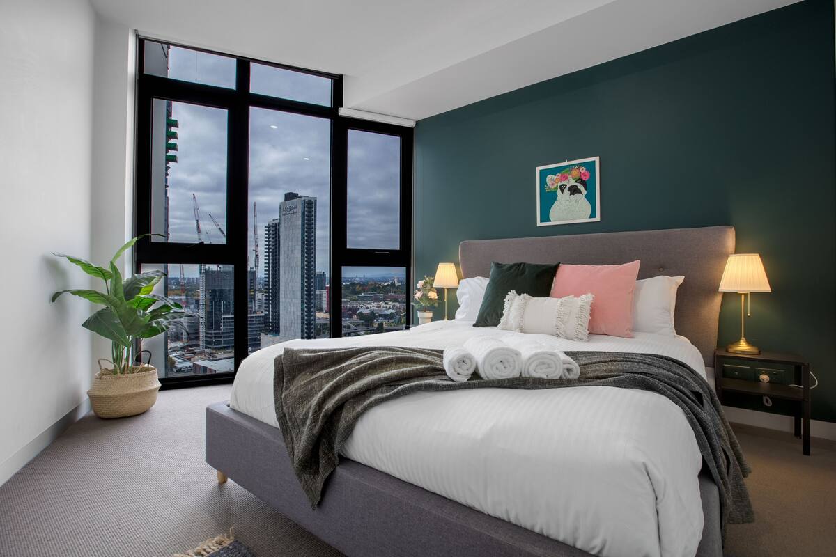 A modern hotel room with a large bed, a view of the cityscape, and a potted plant.