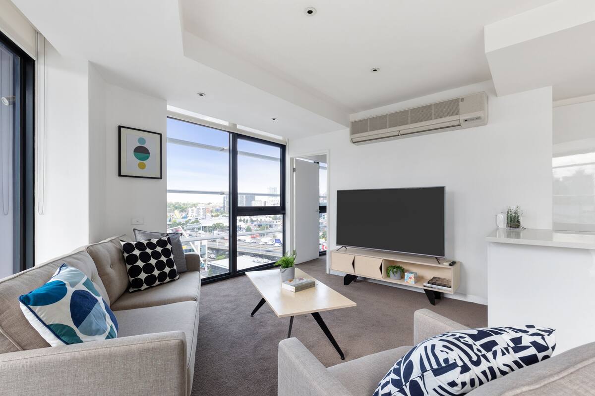 A modern living room with a sofa, a coffee table, a flat-screen TV, and a large window offering a city view.