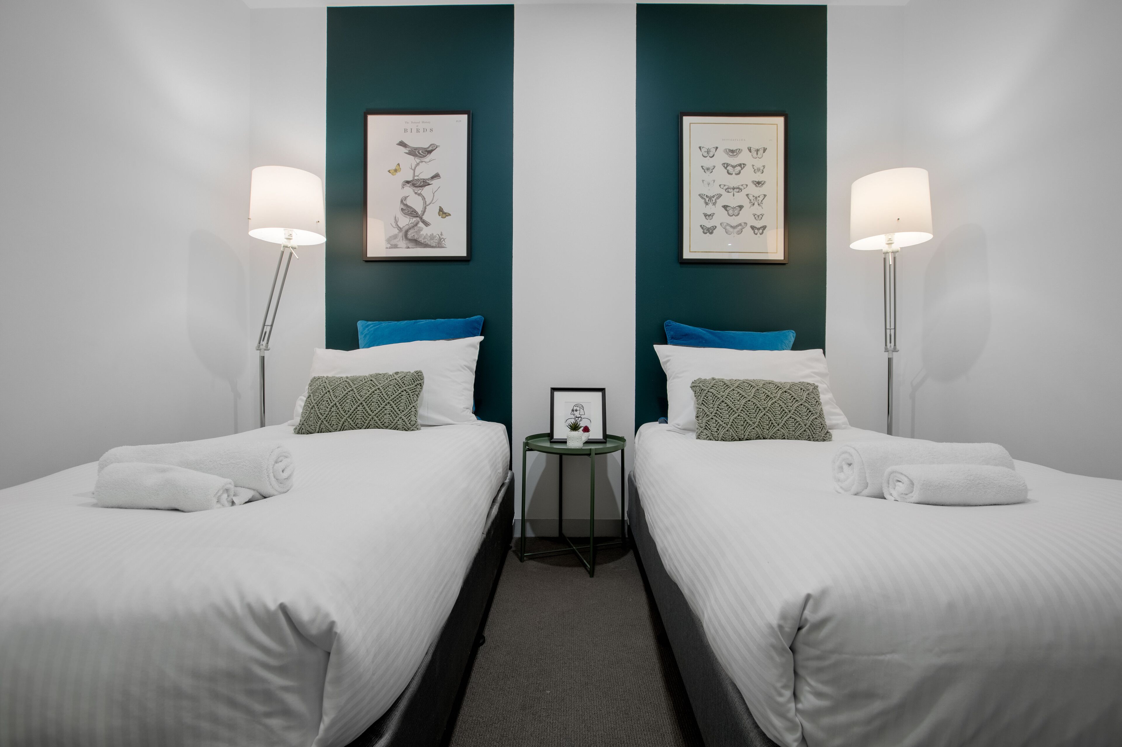 Une chambre d’hôtel avec deux lits simples, une literie blanche, des murs d’accent de couleur turquoise et des œuvres d’art encadrées au-dessus de chaque lit.