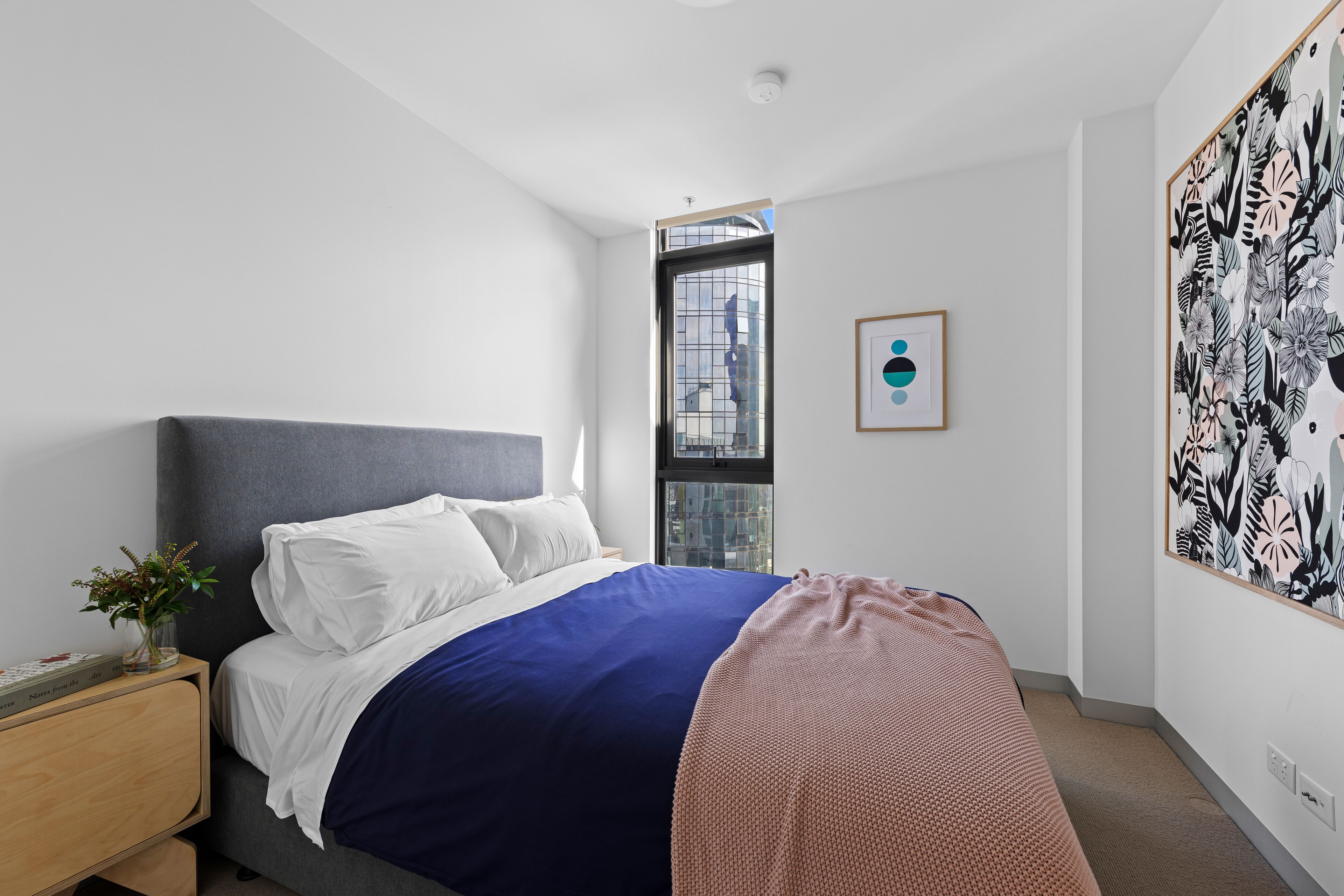 A modern bedroom with a large bed, a nightstand, a window with a view, and a framed artwork on the wall.