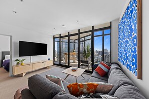 A modern living room with a large TV, a sofa, a coffee table, and a balcony with a city view. - Nook Melbourne Bank Tower Apartments (Southbank)