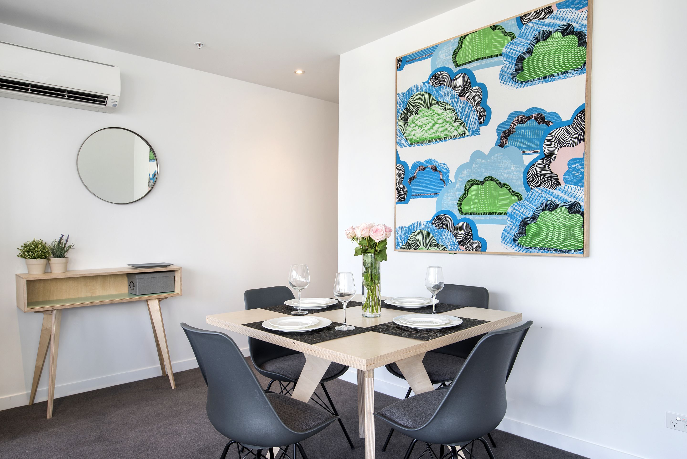 Un coin repas avec une table en bois dressée pour quatre personnes, entourée de chaises noires. Un miroir rond est accroché au mur, à gauche, et une peinture abstraite aux couleurs vives se trouve au-dessus de la table.