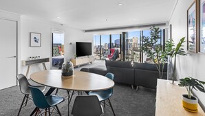 A modern living room with a round dining table, grey and blue chairs, a grey sofa, and a large window with a city view. - Nook Melbourne 173 City Road (Southbank)