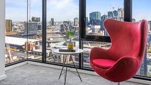 A red chair in a corner of a room with large windows overlooking a cityscape. - Nook Melbourne 173 City Road (Southbank)