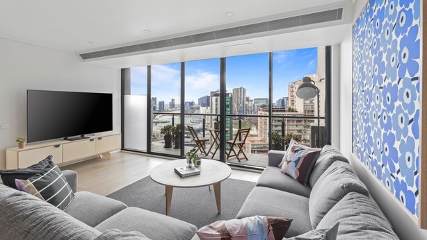 A modern living room with a large TV, comfortable seating, a coffee table, and a balcony with a city view. - Nook Melbourne 173 City Road (Southbank)