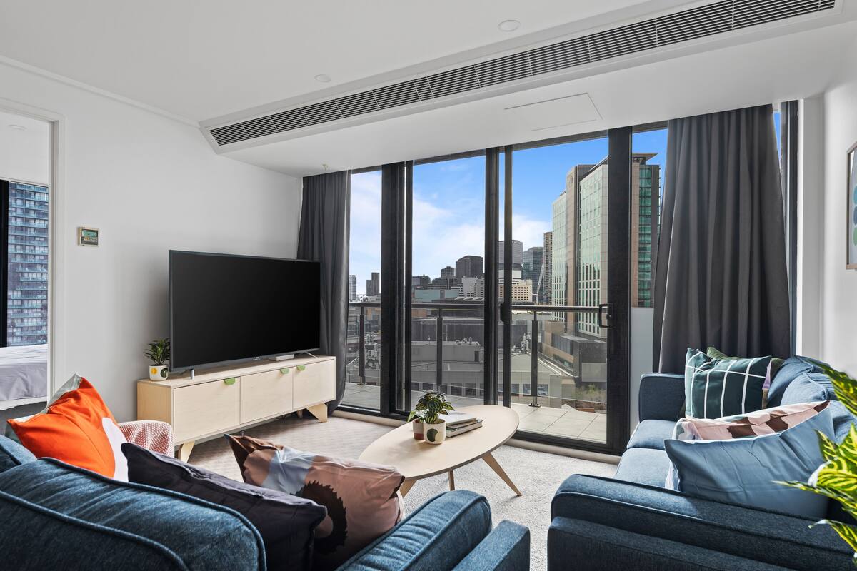 Una sala de estar moderna con un televisor de pantalla plana grande, un sofá, una mesa de centro y vistas a la ciudad a través de amplias ventanas.