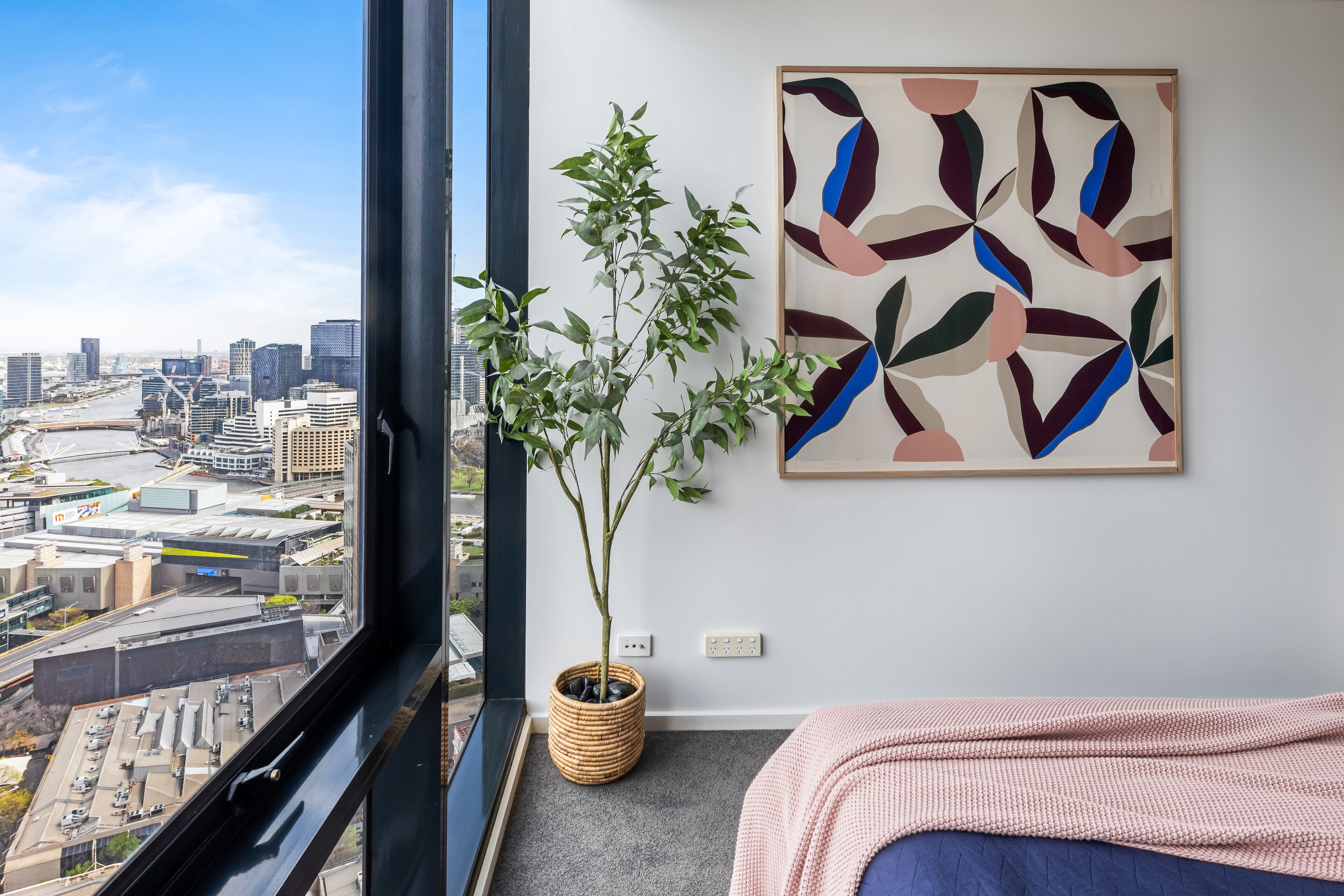 A bedroom with a city view, a potted plant, and a modern abstract painting.
