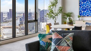 A modern living room with a large window overlooking a cityscape, a round dining table, and a sofa with colorful cushions. - Nook Melbourne 173 City Road (Southbank)