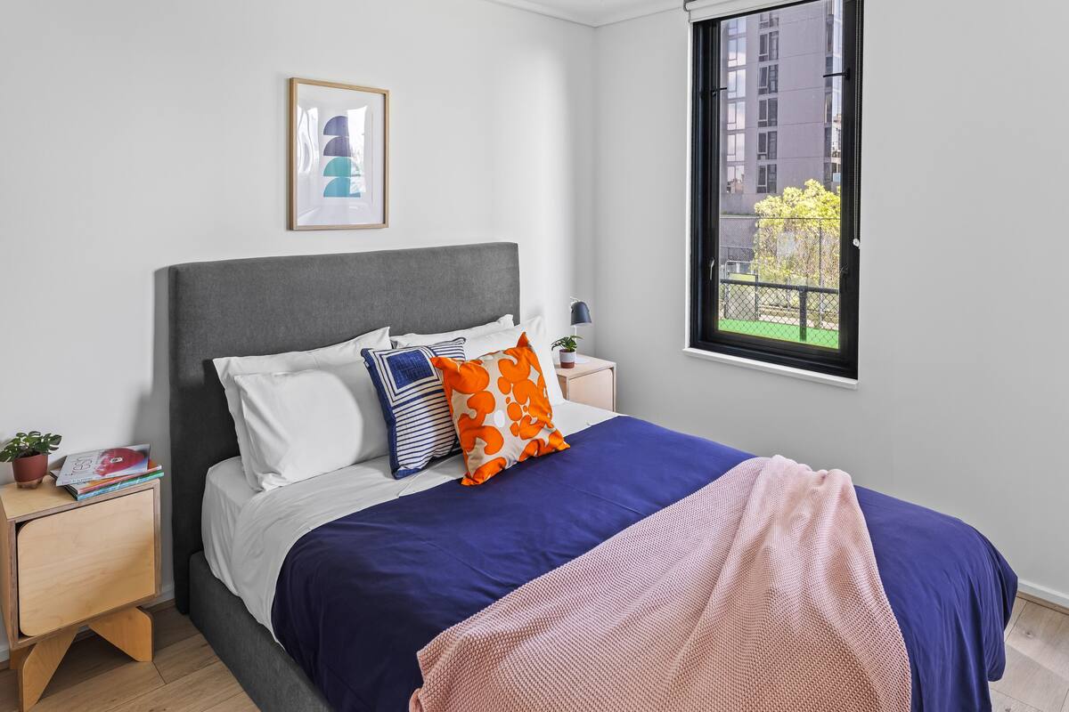 Un dormitorio ordenado con cama, almohadas, una mesita de noche y una ventana. En la pared, se aprecia un cuadro enmarcado.