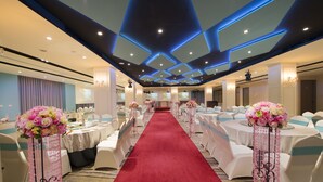 Un salón de hotel con alfombra roja, mesas y sillas blancas, y centros de mesa florales.