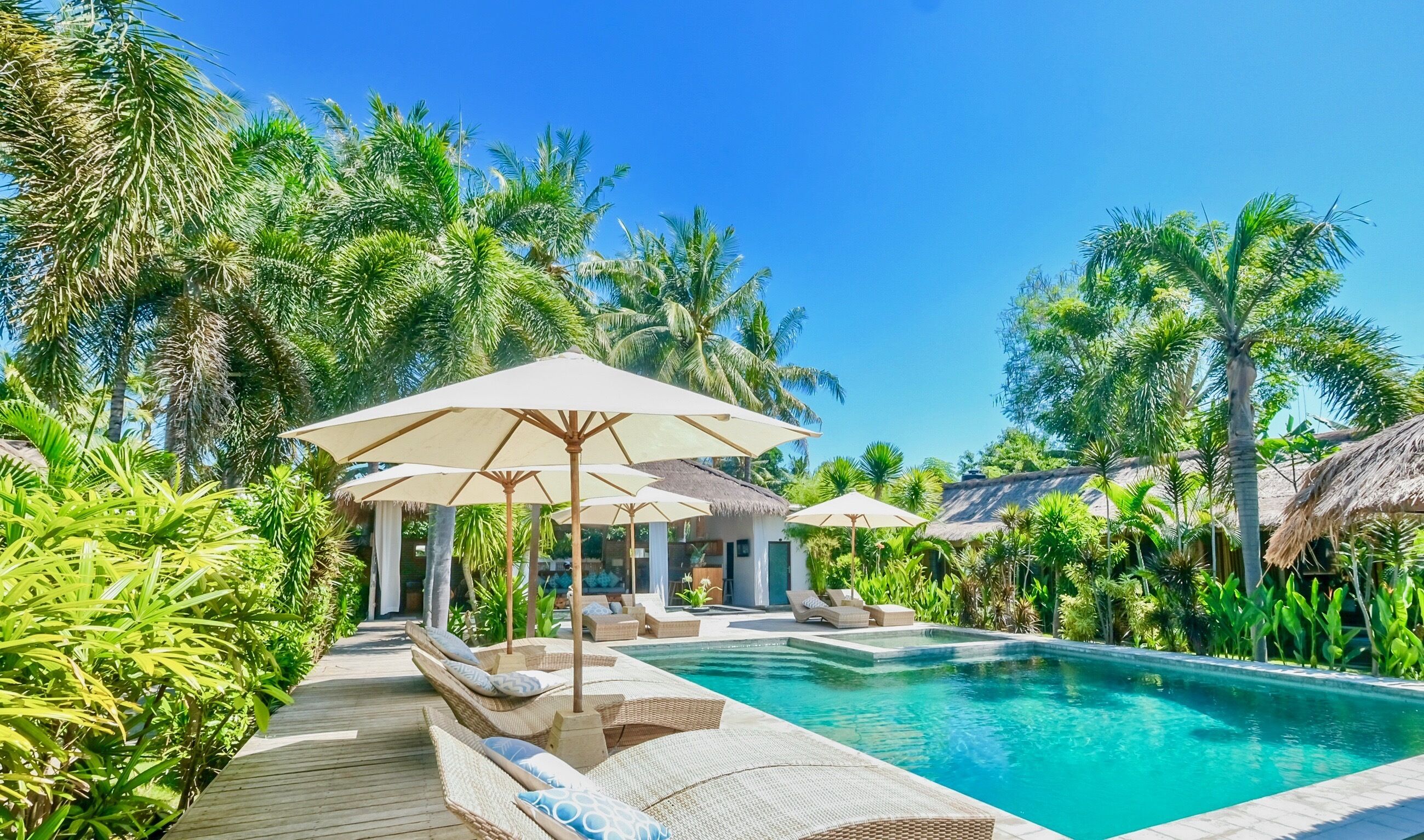 Zona de piscina con sillones de descanso y sombrillas, rodeada de exuberante vegetación y palmeras.