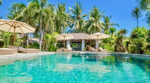Una piscina cristalina con sillones de descanso y sombrillas, rodeada de palmeras y una estructura con techo de paja.