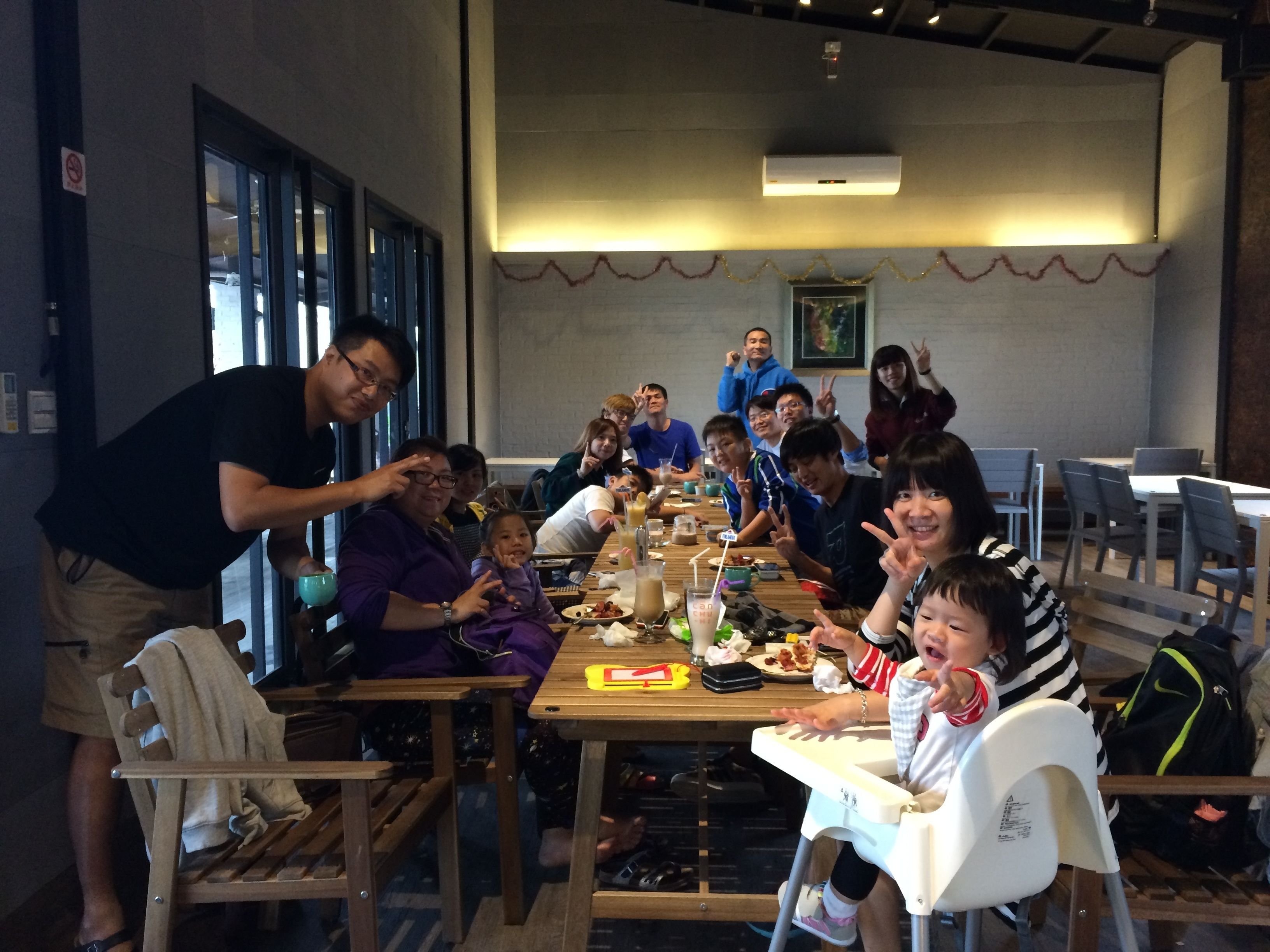 A group of people seated around a table in a dining area with a high ceiling and a large window.