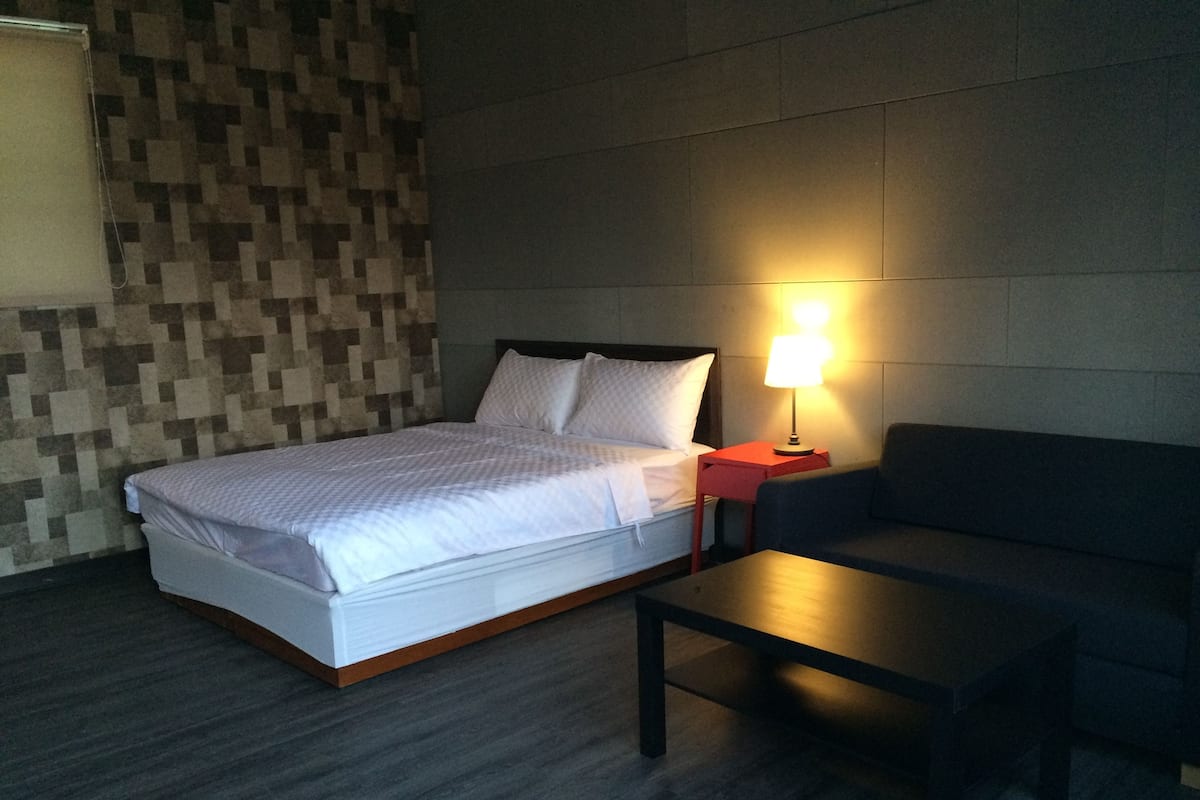 Una habitación de hotel con una cama, una mesita de noche con lámpara, un sofá y suelo de madera.