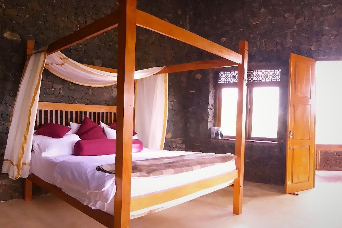 A four-poster bed with white and pink bedding, a stone wall background, and a wooden door.