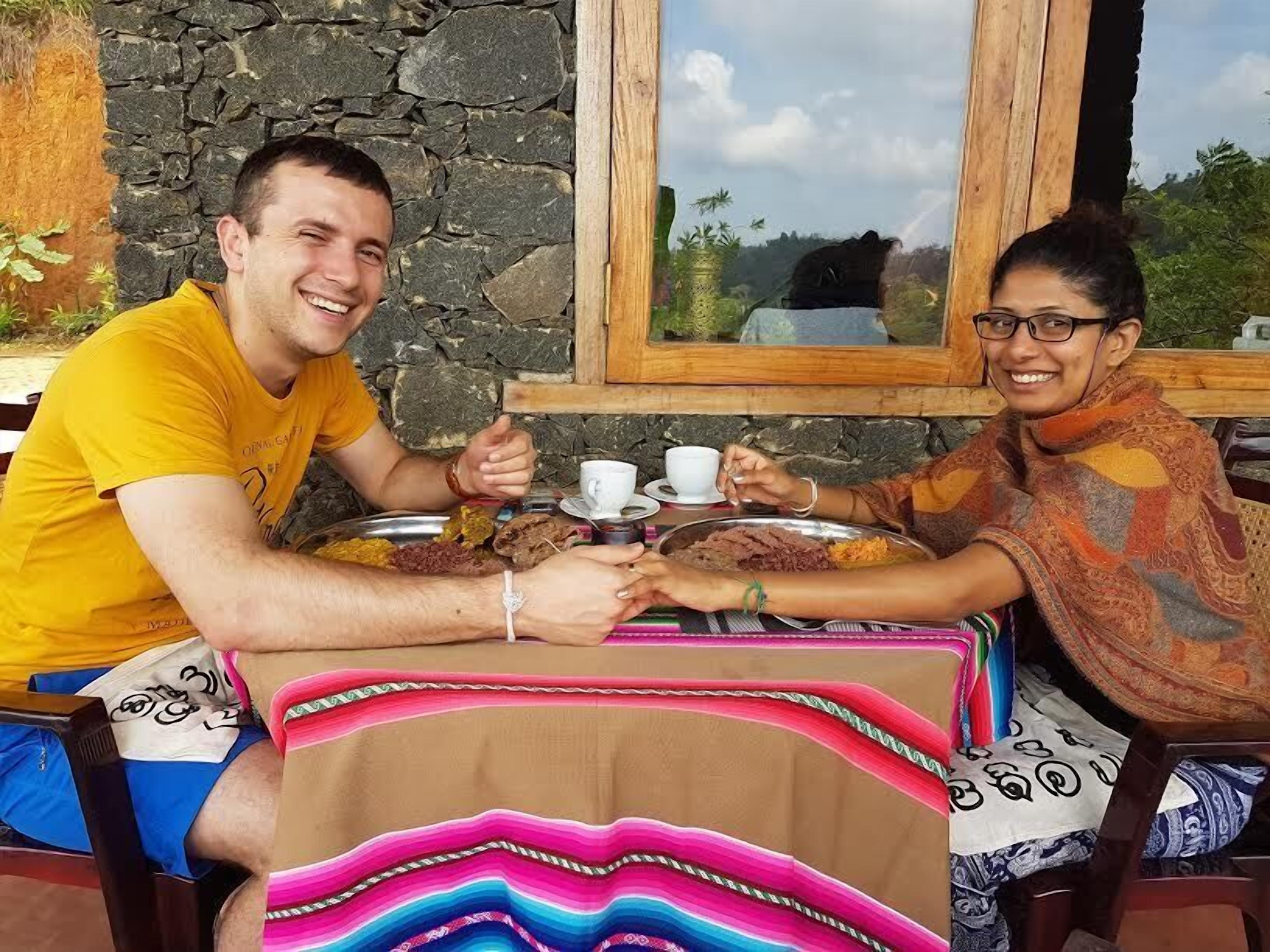 Two people sitting at a table with a stone wall behind them, enjoying a meal.