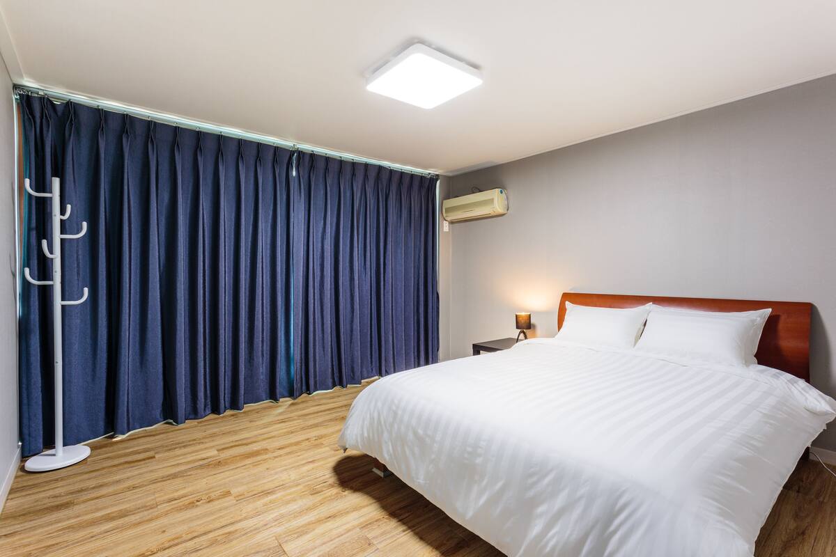 A hotel room with a bed, wooden flooring, a ceiling light, an air conditioning unit, and a nightstand with a lamp.