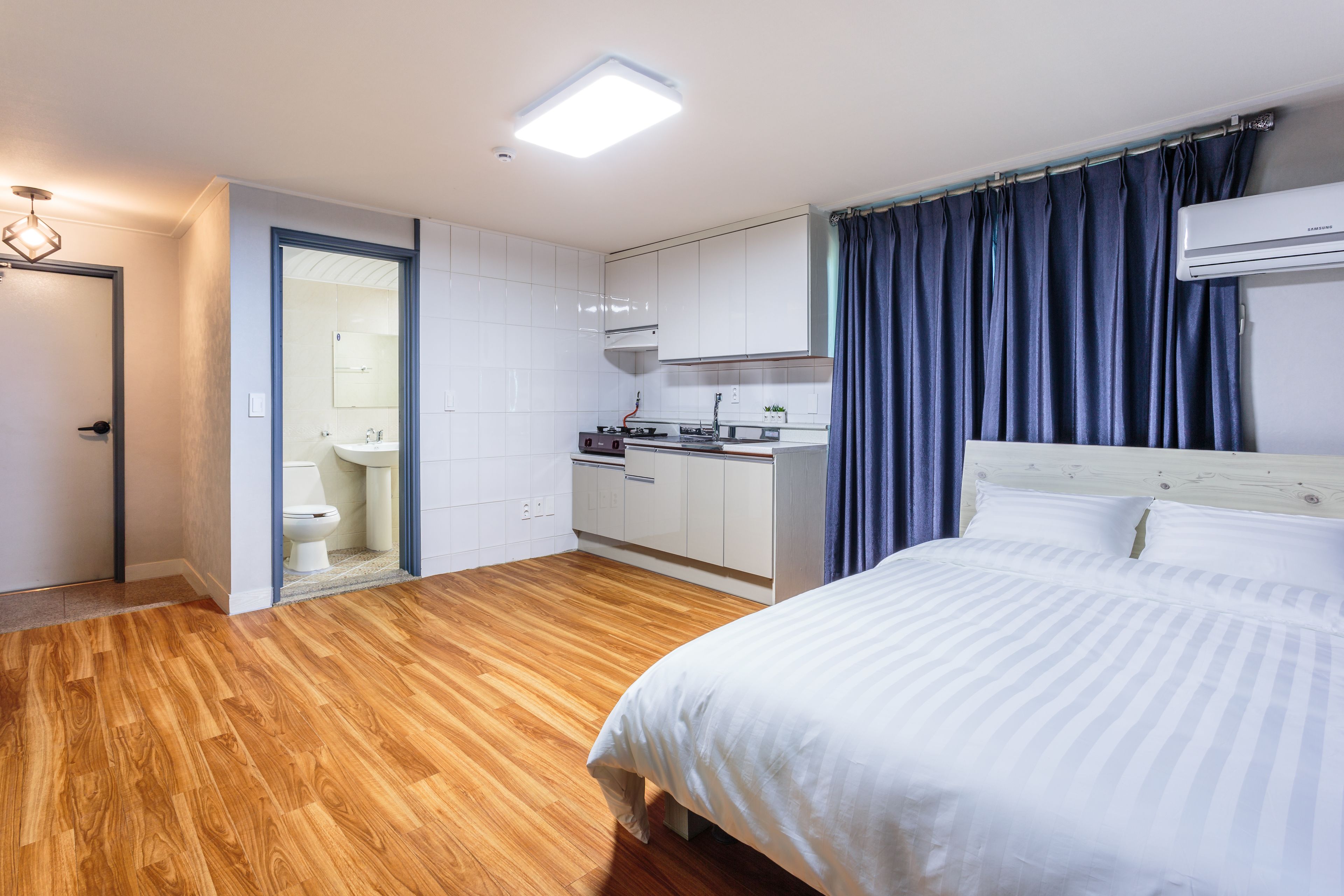 Una moderna camera d'albergo con pavimento in legno, un letto con biancheria bianca, angolo cottura e bagno con WC e lavandino.