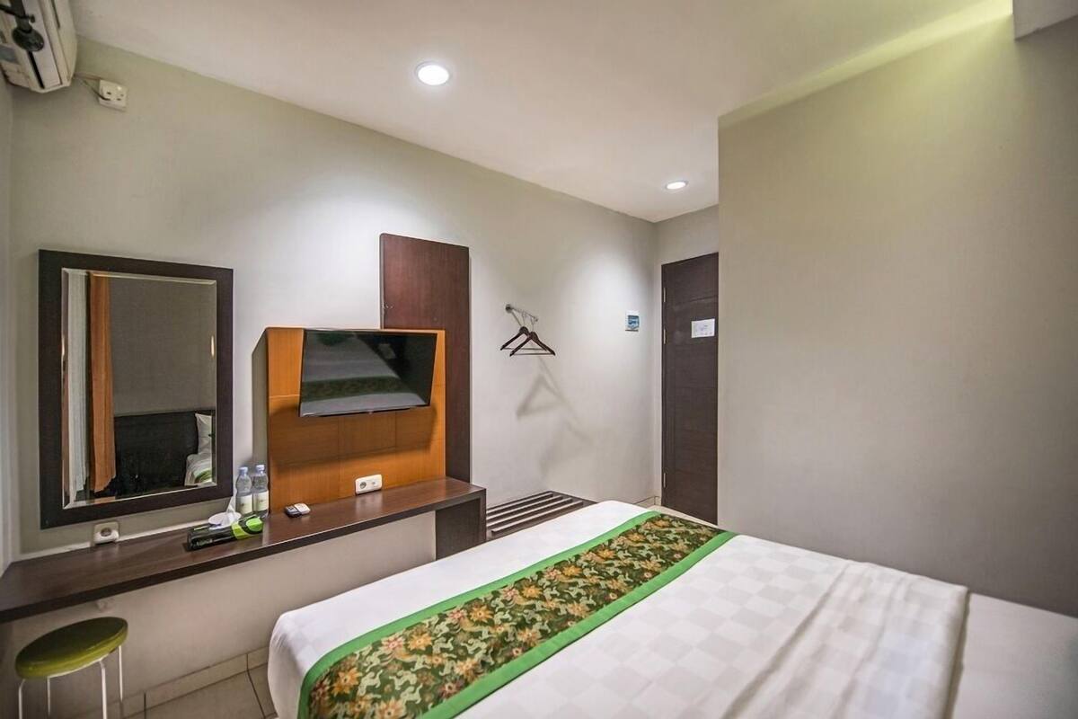 A hotel room with a bed, a desk with a mirror, a TV, a hanging coat rack, and a green-striped bed runner.