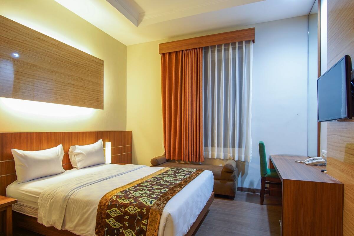A hotel room with a bed, a desk, a chair, a TV, and a window with curtains.