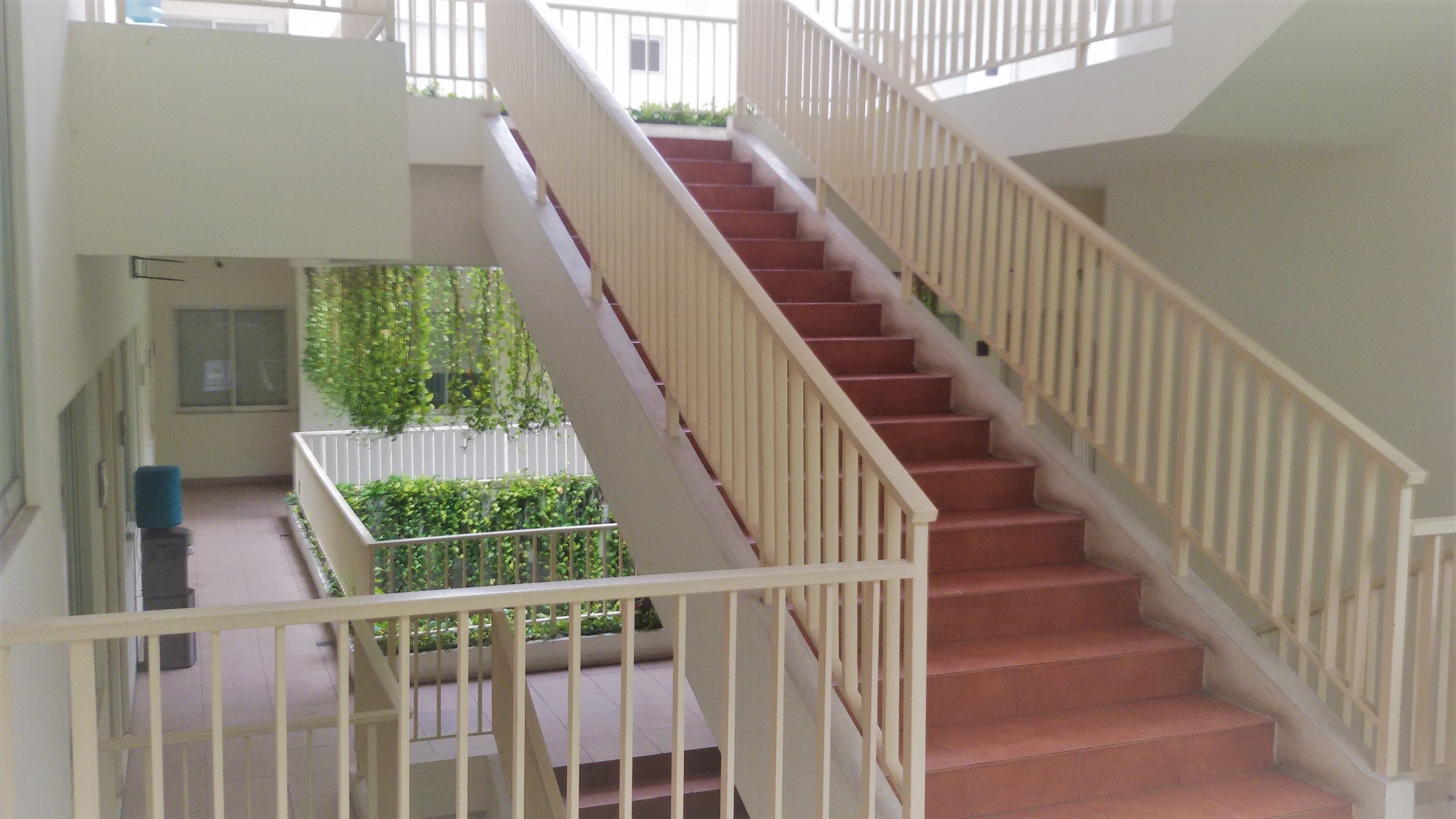 Un interior de varios niveles con una escalera, un balcón con vegetación y una barandilla blanca.