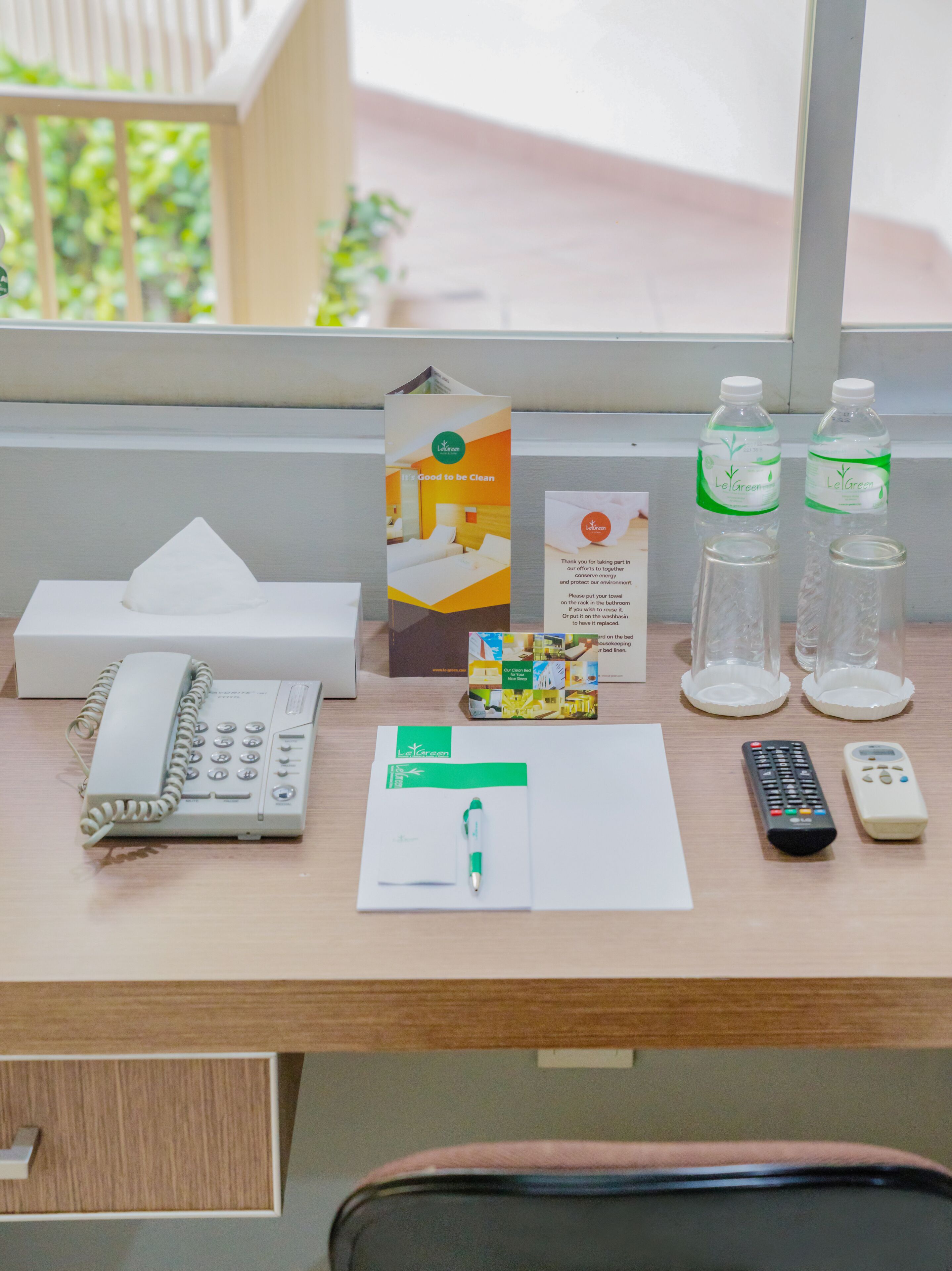 A hotel desk with a telephone, remote controls, a notepad, a pen, a tissue box, a brochure, and two water bottles.