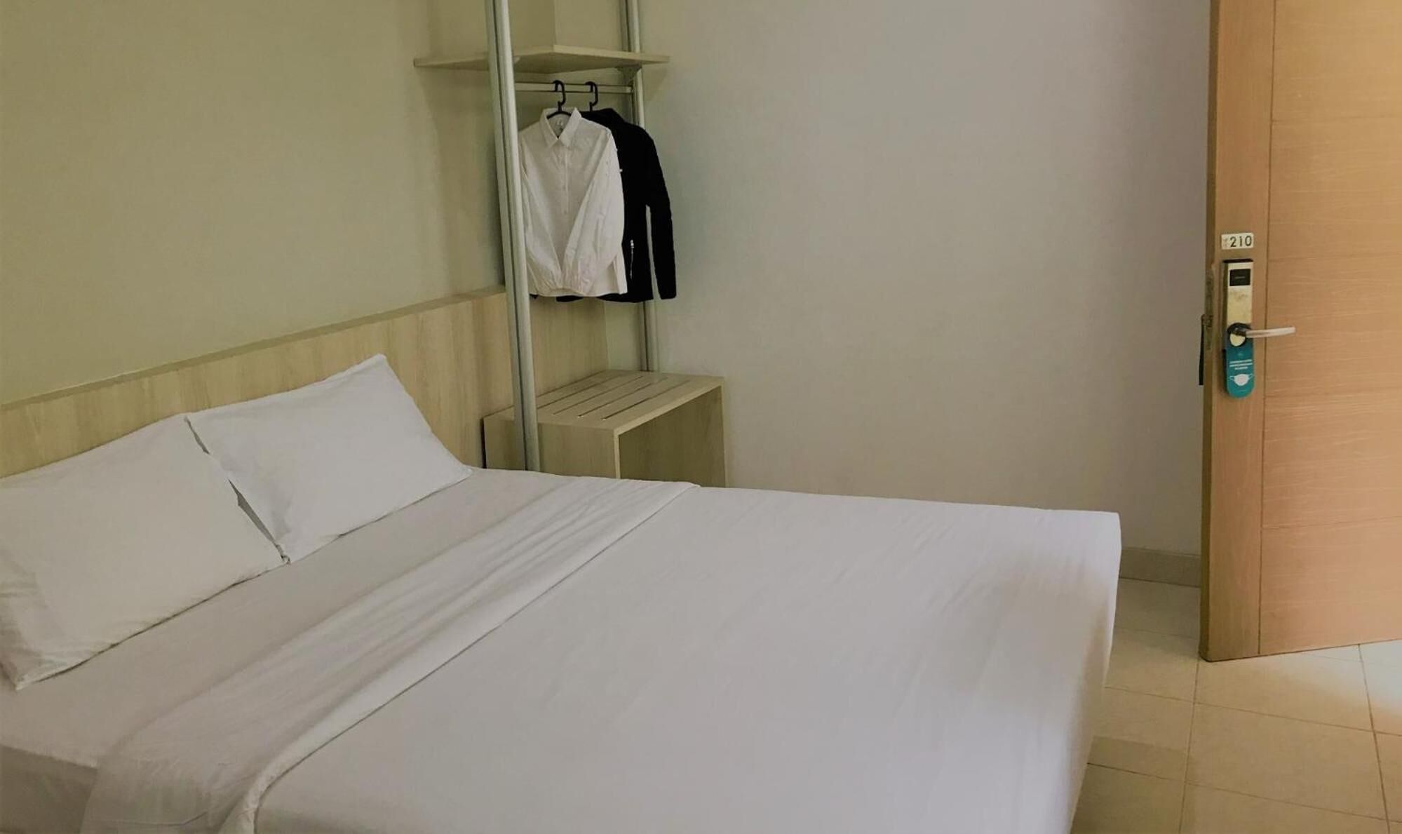 A hotel room with a bed, a small wooden nightstand, a clothing rack, and a door with a lock.