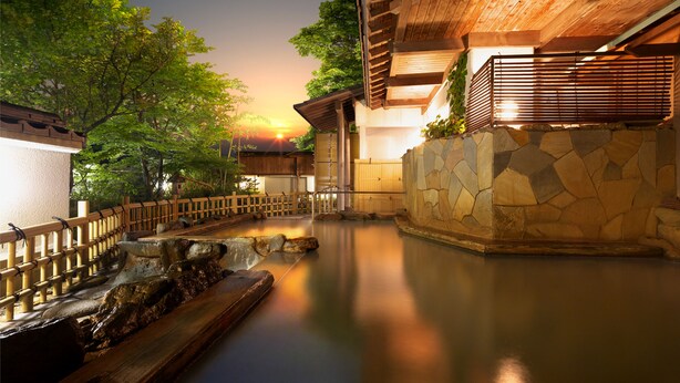 A serene outdoor pool area with a wooden deck and stone walls, surrounded by lush greenery and a sunset in the background. - Moriaki Ryokan (Shibukawa)