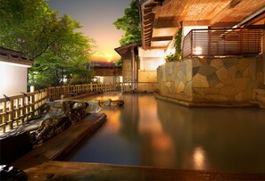 Área tranquila de piscina al aire libre con muelle de madera y paredes de piedra, rodeada de exuberante vegetación y un atardecer al fondo.