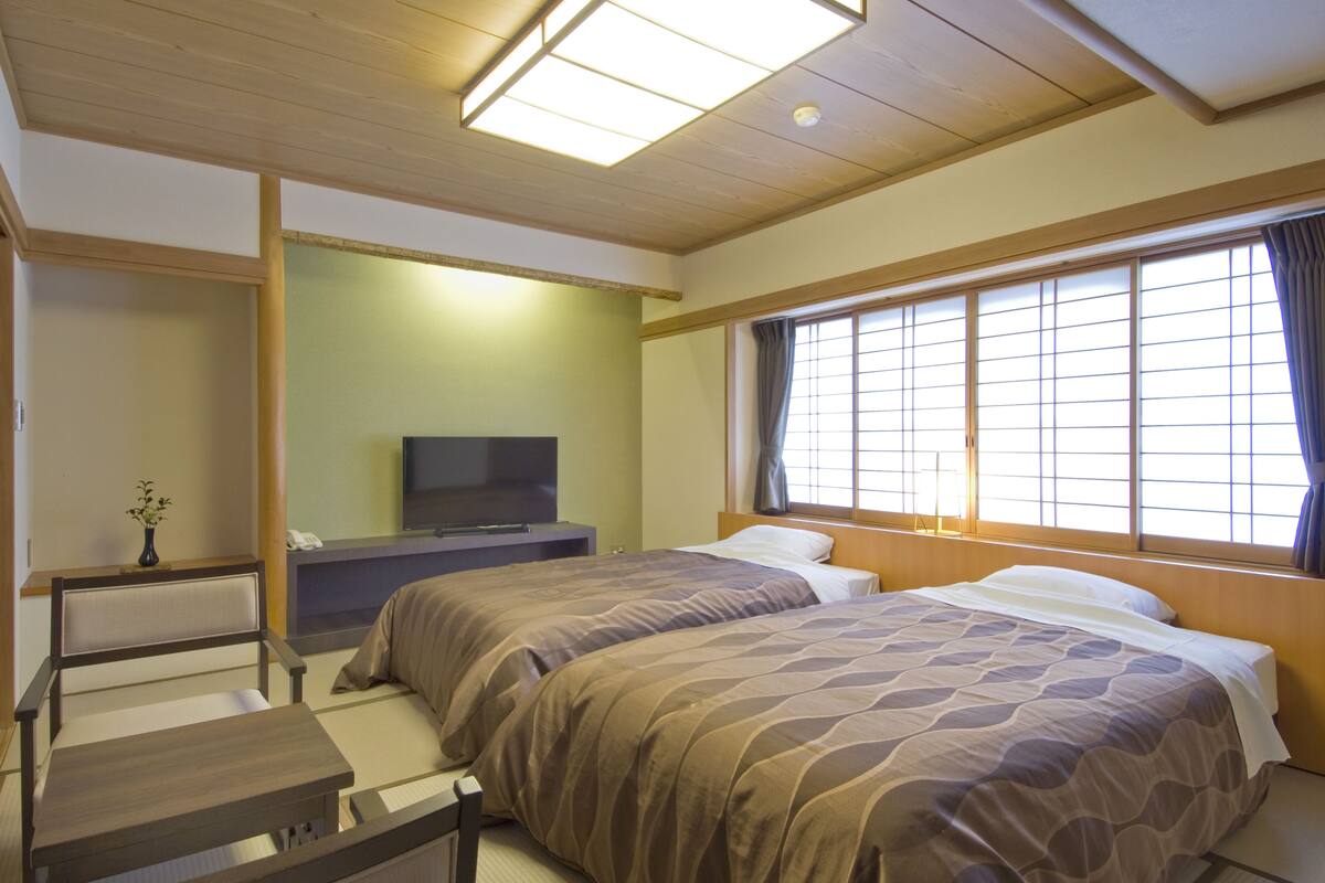 A hotel room with two beds, a TV, a desk, and a window with curtains.